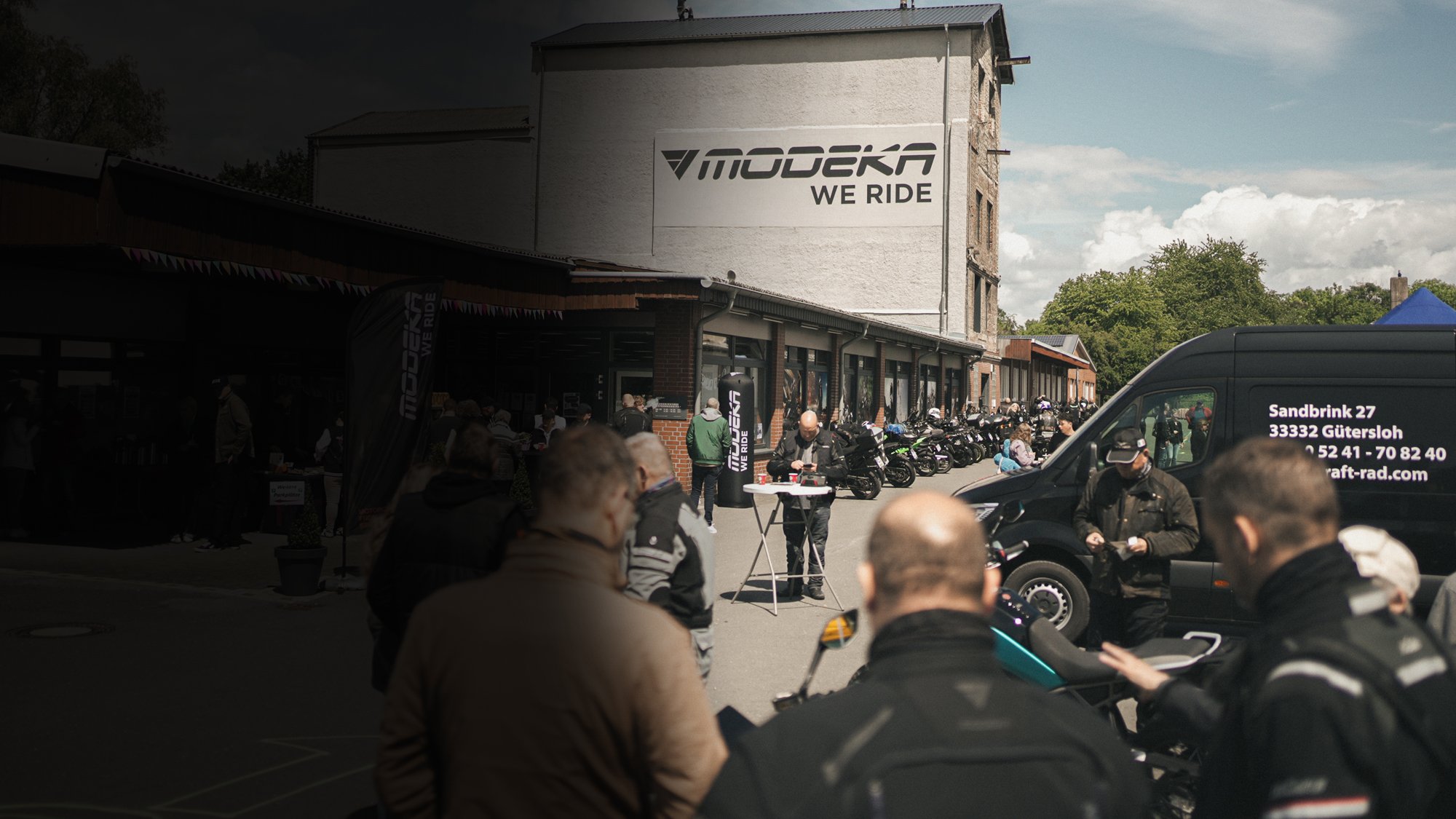 Besucher und Motorräder auf dem Gelände eines Motorrad-Events vor einem Gebäude mit Modeka-Logo.