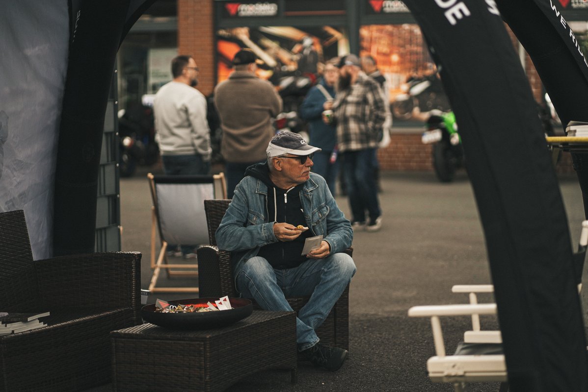 Mann sitzt bei einem Motorrad-Event in einer Lounge und isst, während sich im Hintergrund Besucher unterhalten.