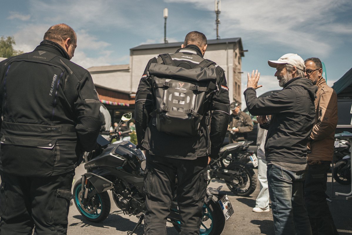 Besucher stehen bei einem Motorrad-Event zwischen geparkten Maschinen und unterhalten sich auf dem Außengelände