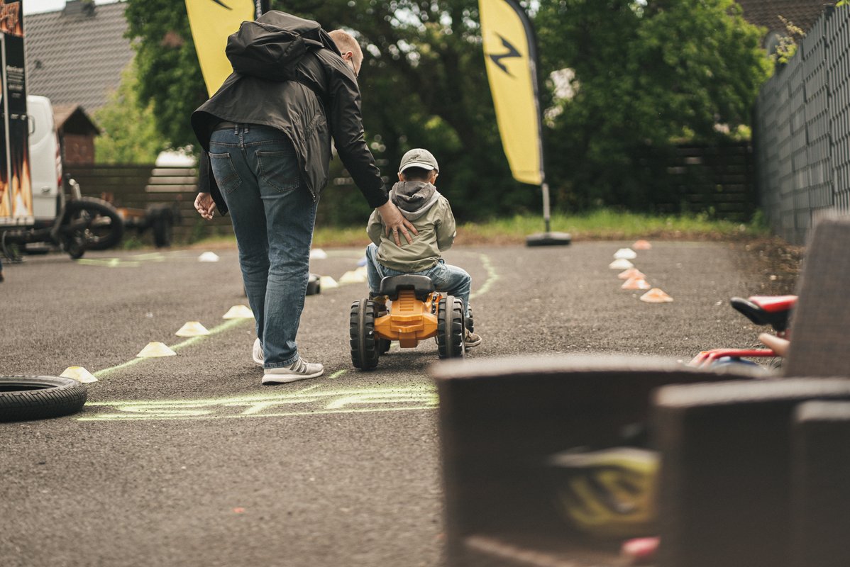 Kind fährt auf einem kleinen Fahrzeug durch einen markierten Parcours, während eine erwachsene Person unterstützend daneben geht.