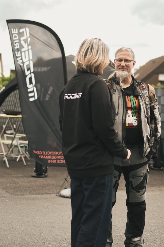 Zwei Personen unterhalten sich vor einer Modeka-Flagge auf einem Motorrad-Event.