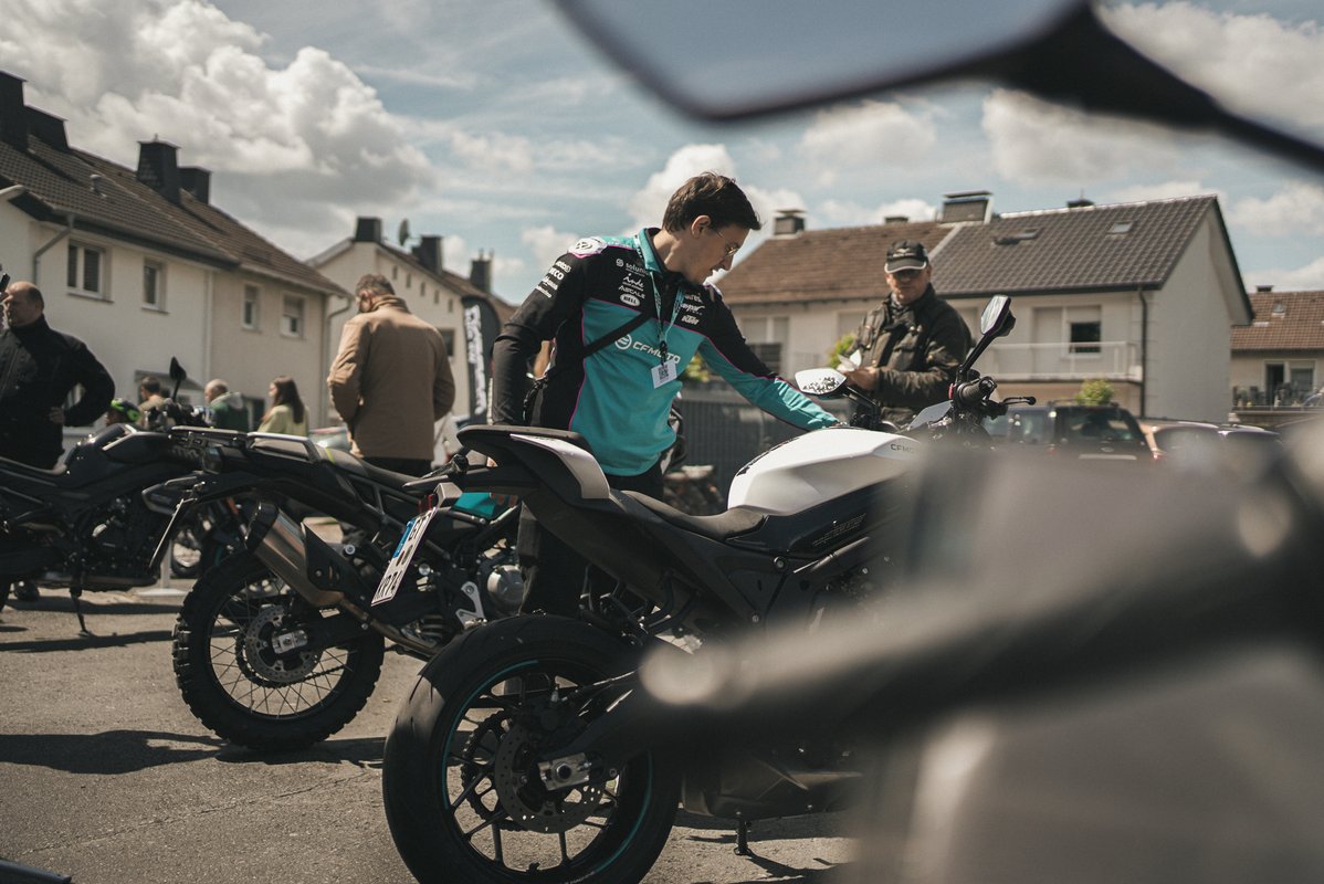 Person in CF Moto-Kleidung sitzt auf einem weißen Motorrad, während Besucher im Hintergrund weitere Motorräder ansehen.