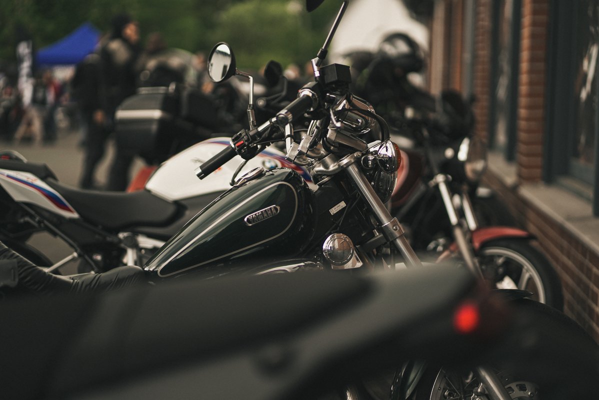 Nahaufnahme mehrerer geparkter Motorräder bei einem Event, im Vordergrund ein dunkles Motorrad mit Fokus auf Tank und Lenker.
